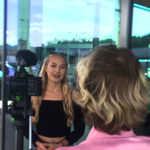 Ein Videojournalist dreht ein Interview mit einer blonden Frau die vor einer Glaswand steht. Blick über die Schulter des Videosjournalisten