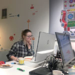 Eine junge Frau in einer karierten Bluse sitzt an einem Schreibtisch und arbeitet am Computer.